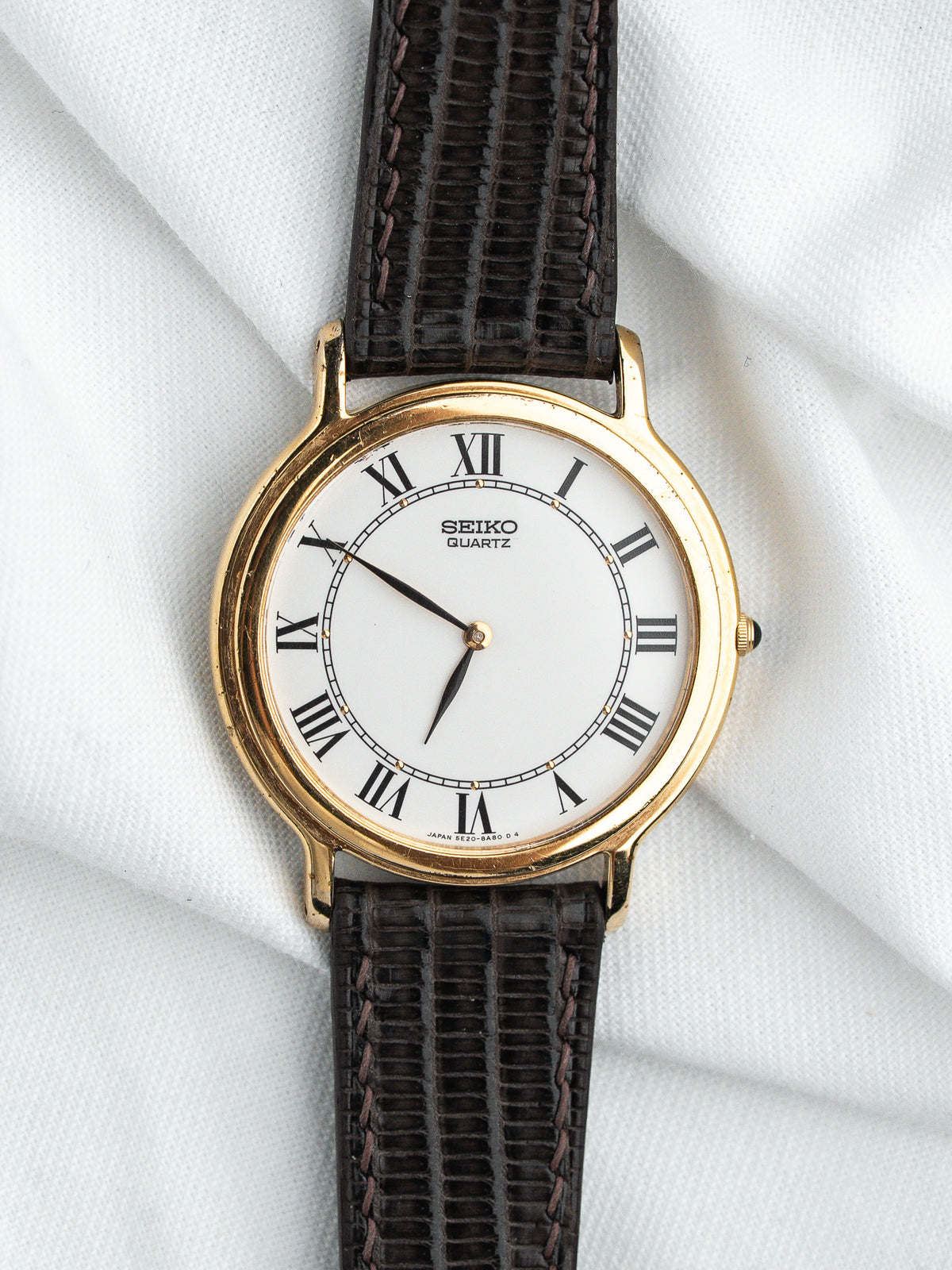 Montre Seiko Quartz vintage au boîtier doré, cadran blanc et bracelet en cuir marron.