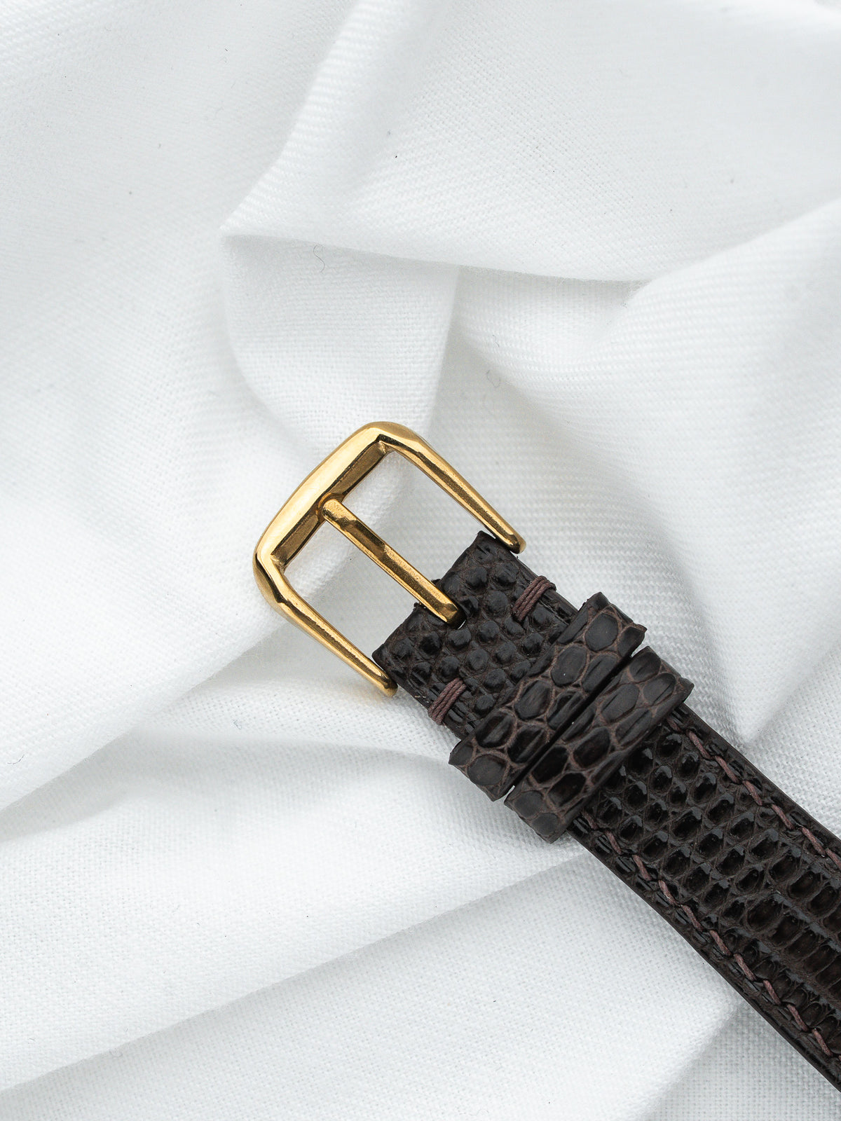 Détail d'un bracelet de montre en cuir brun texturé avec boucle dorée sur tissu blanc.