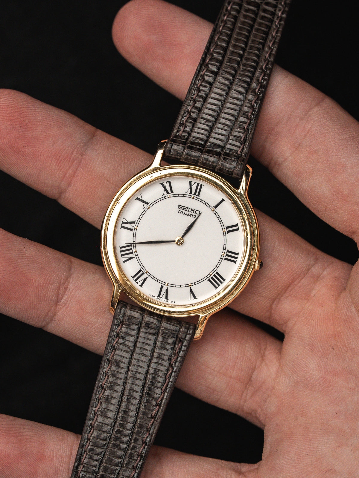 Montre Seiko Quartz vintage avec cadran blanc, chiffres romains et bracelet en cuir gris.