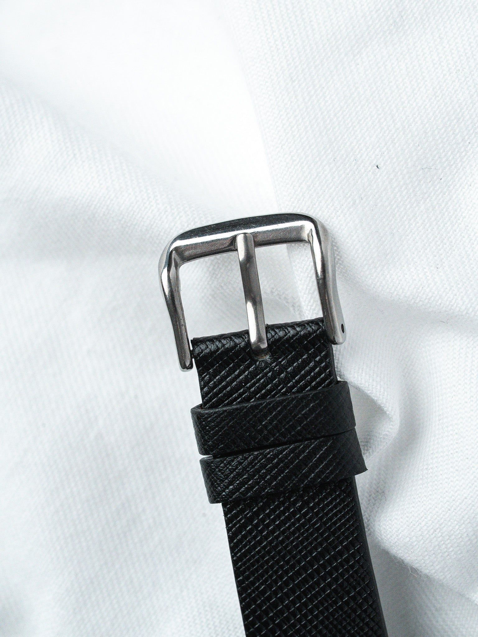 Black textured leather watch strap with polished steel buckle on white fabric