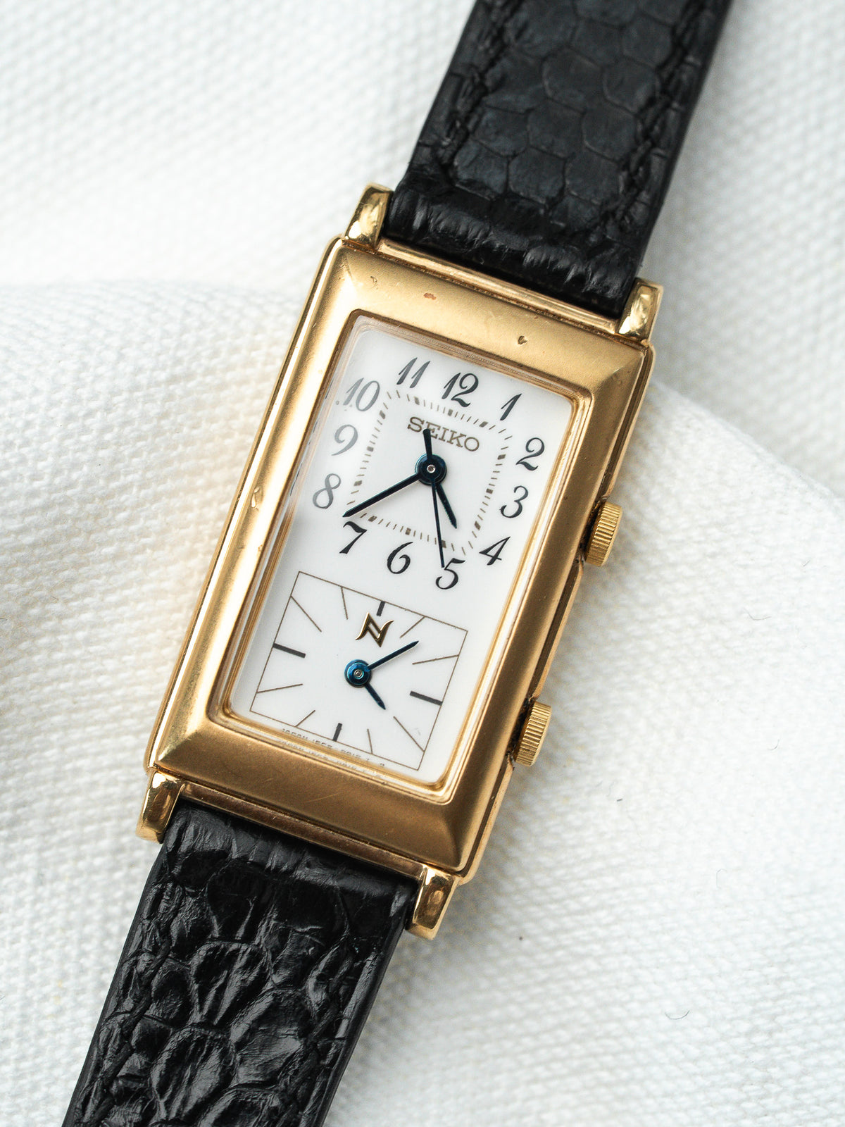 Montre Seiko vintage rectangulaire en or avec bracelet en cuir noir et cadran blanc.