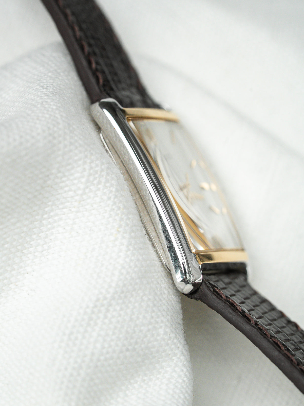 Montre vintage rectangulaire avec boîtier argenté et bracelet en cuir brun sur tissu blanc.