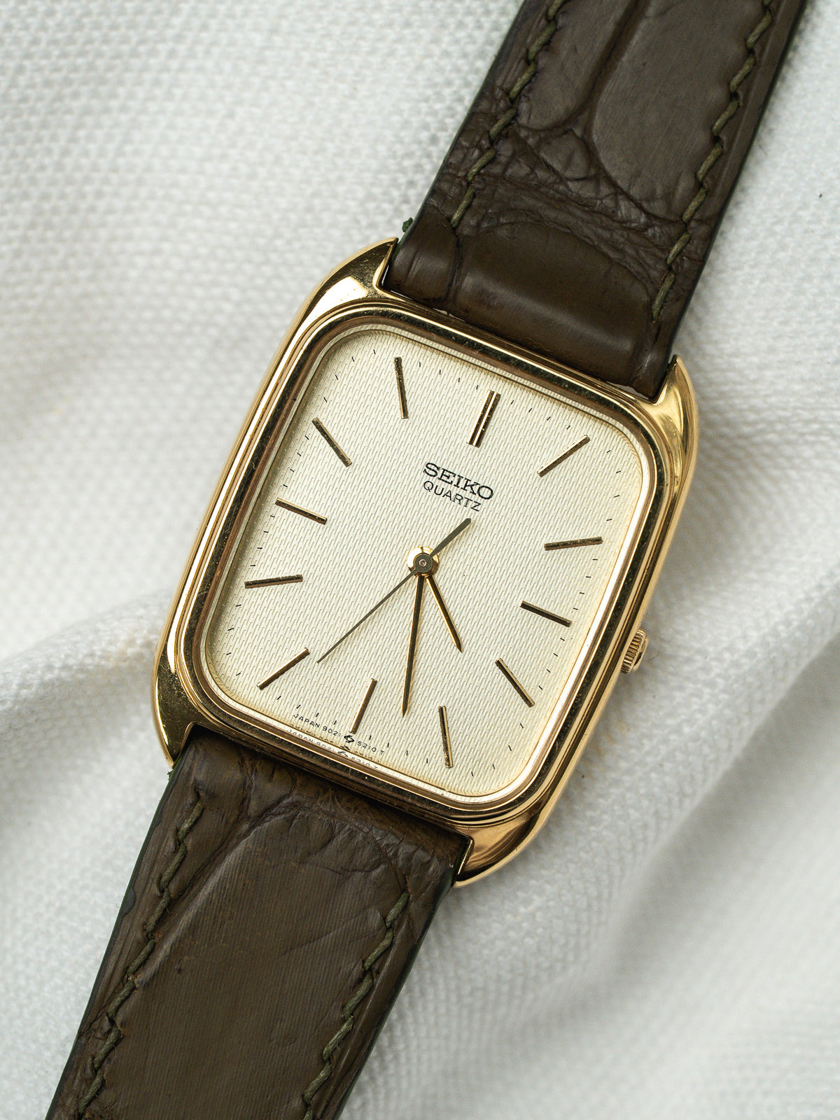 Montre Seiko Quartz vintage au boîtier doré rectangulaire et bracelet en cuir marron.