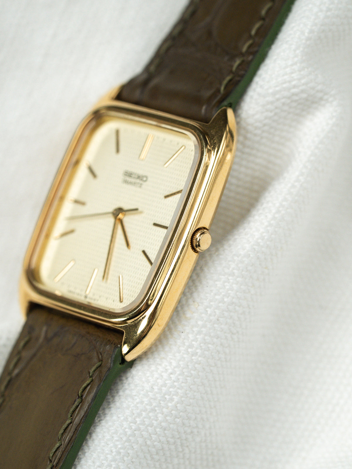 Montre vintage au boîtier rectangulaire doré avec cadran blanc et bracelet en cuir brun.