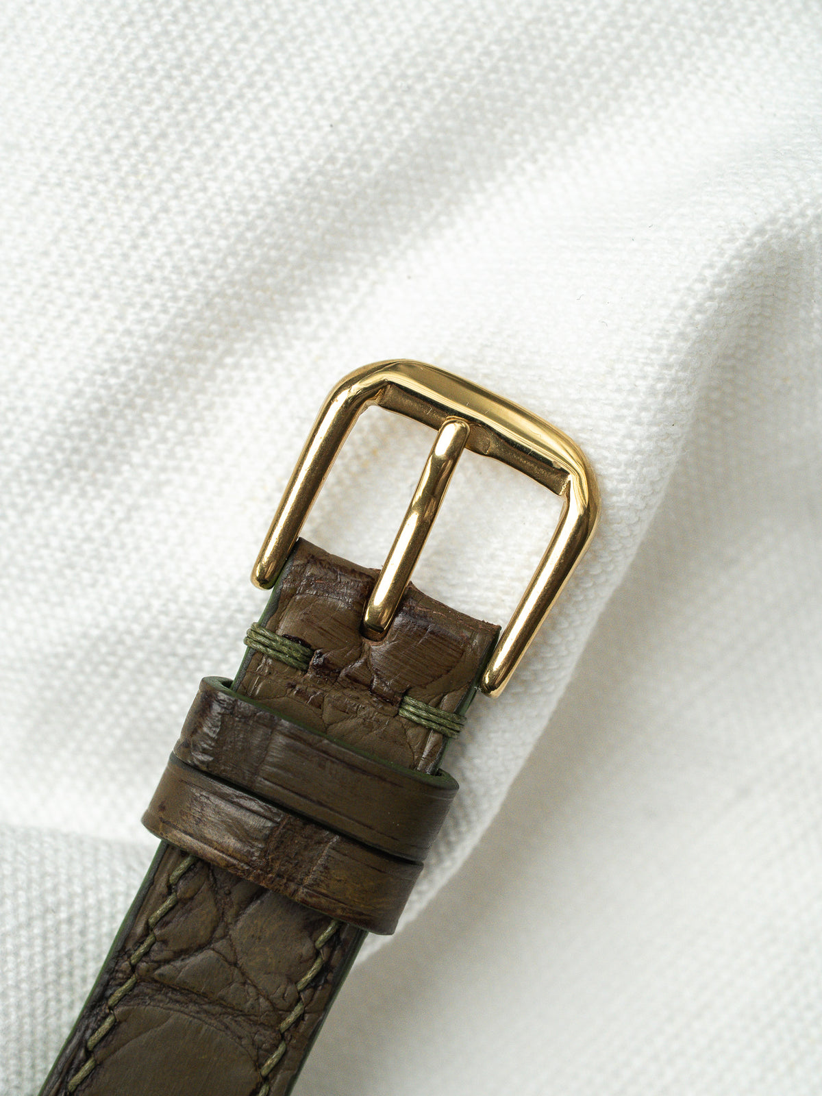 Boucle dorée et bracelet en cuir marron texturé sur un fond textile blanc.