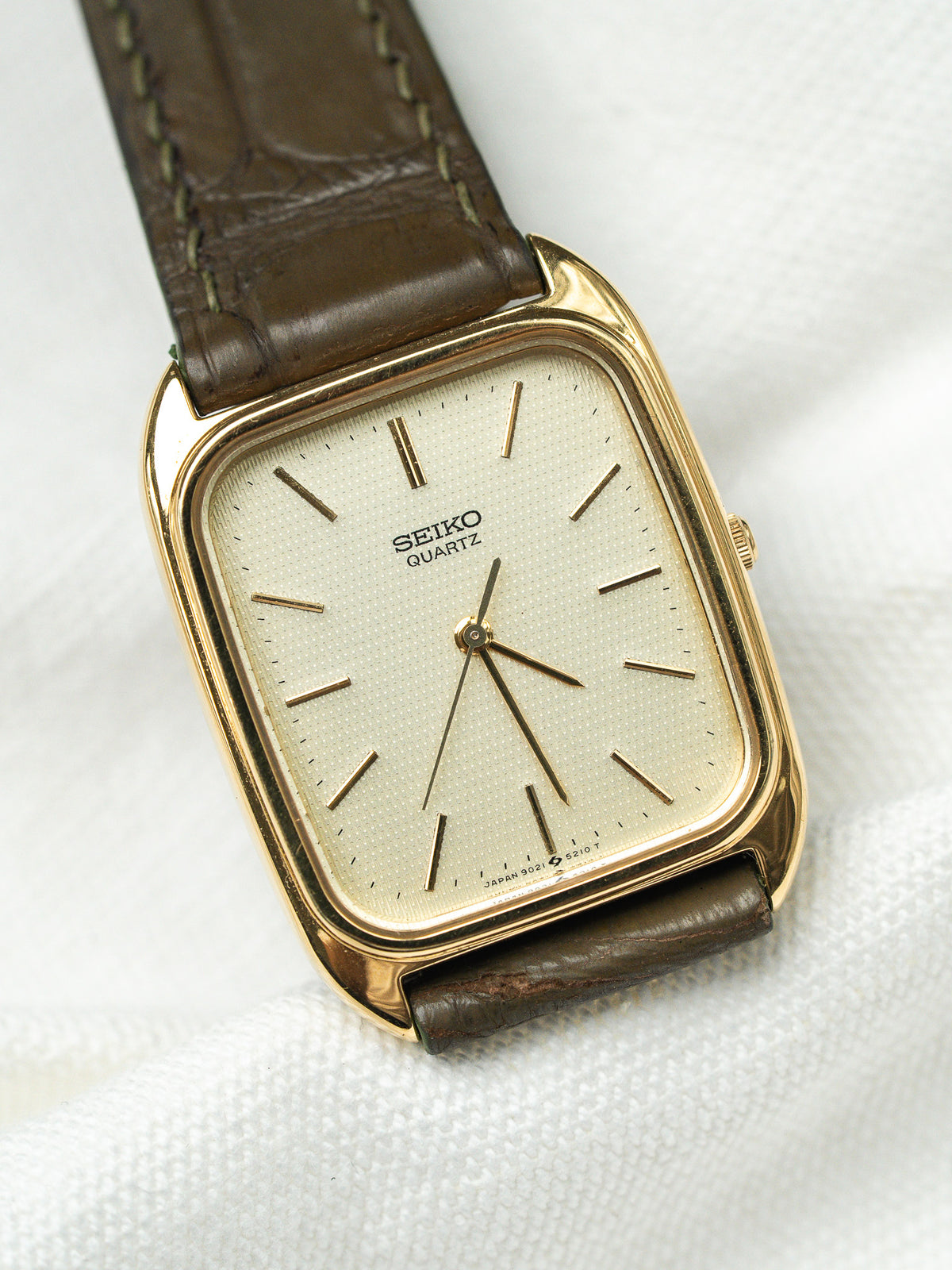 Montre Seiko Quartz vintage au boîtier rectangulaire doré et bracelet en cuir marron.