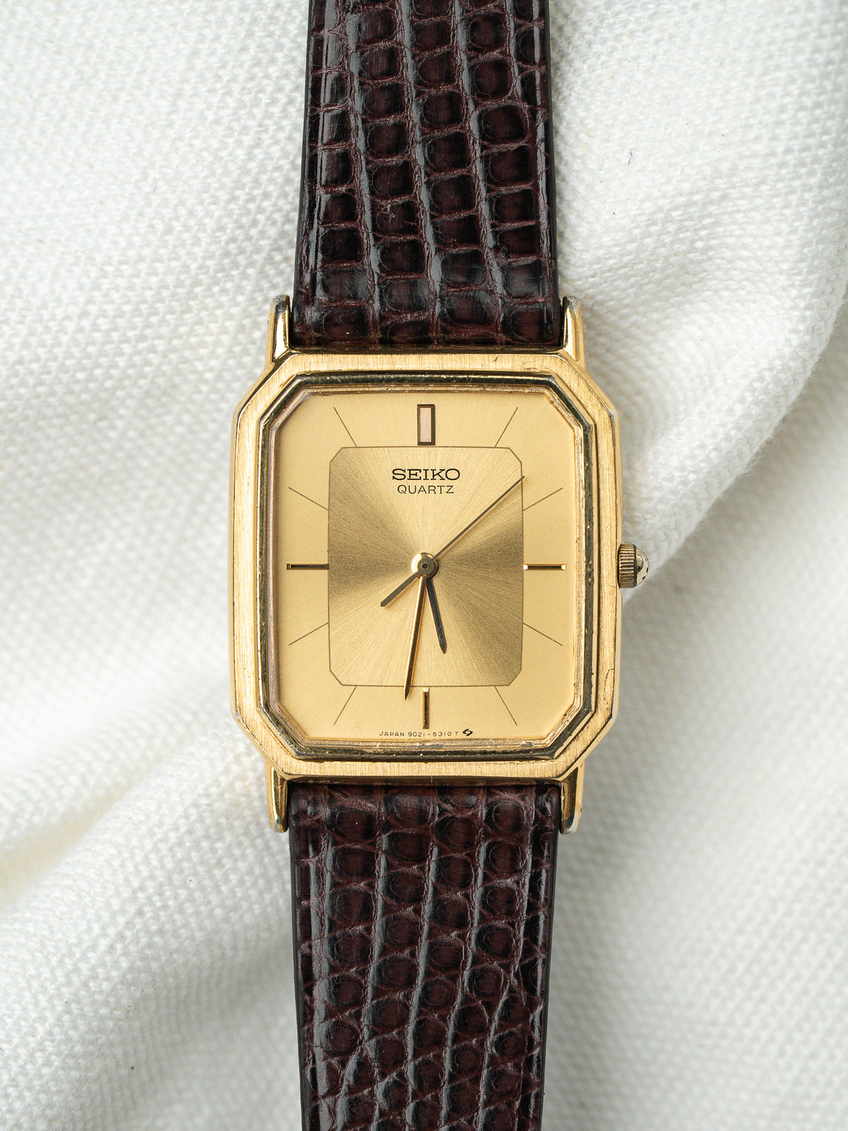 Montre Seiko Quartz vintage au boîtier doré rectangulaire et bracelet en cuir marron.