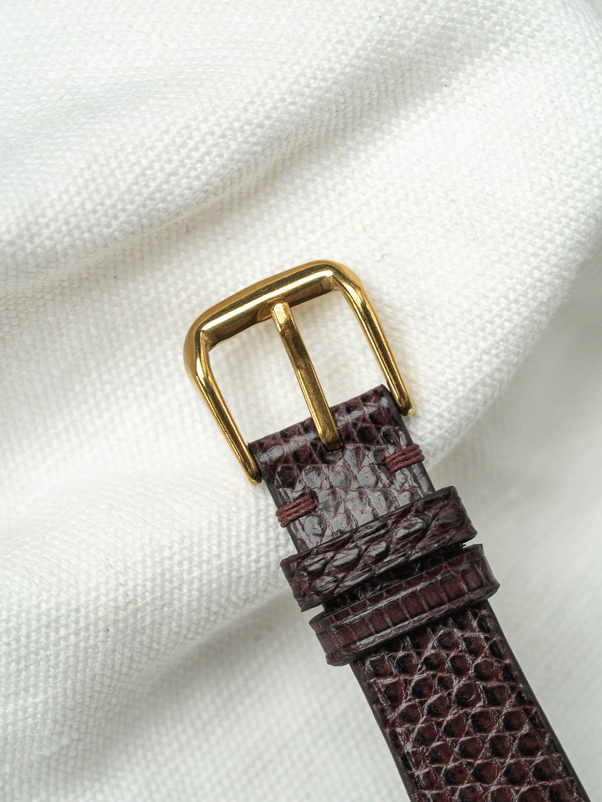 Boucle dorée et bracelet en cuir marron grainé sur un tissu blanc.