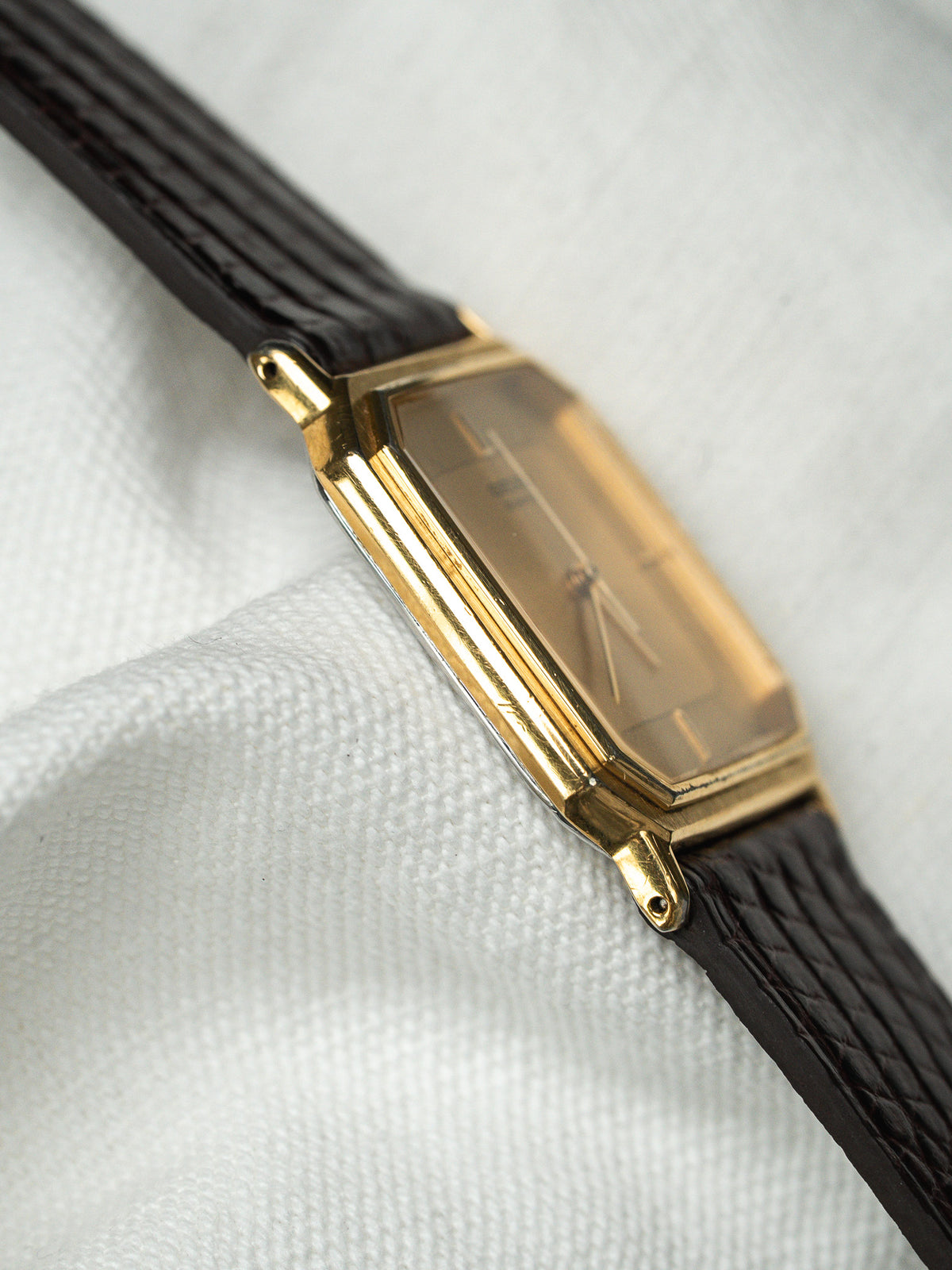 Montre vintage rectangulaire en or avec bracelet en cuir noir sur fond blanc.