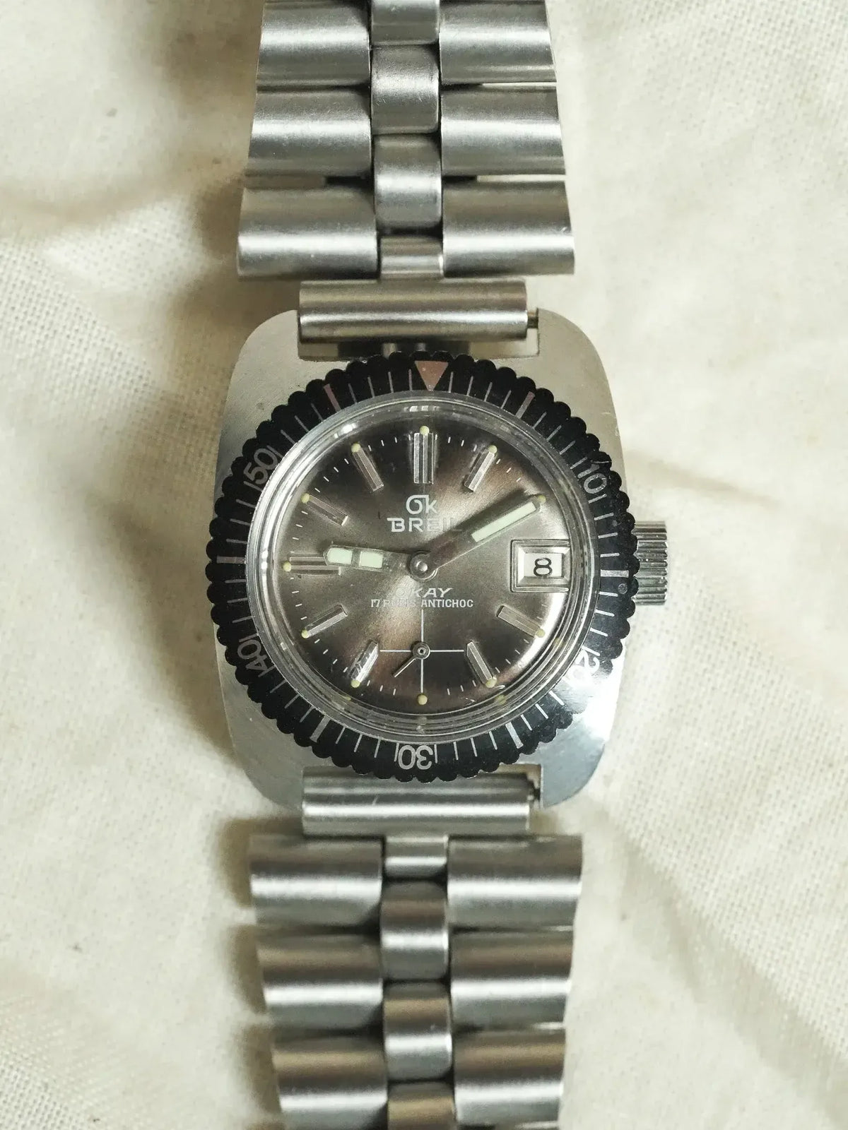 Vintage Breil OKAY watch with black bezel, grey dial, and steel bracelet on fabric