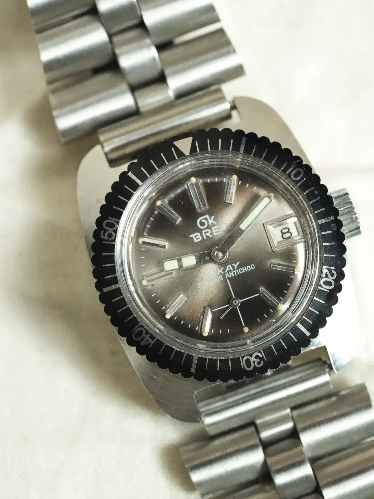 Vintage Breitling Okay watch with black bezel, grey dial, steel bracelet on neutral background