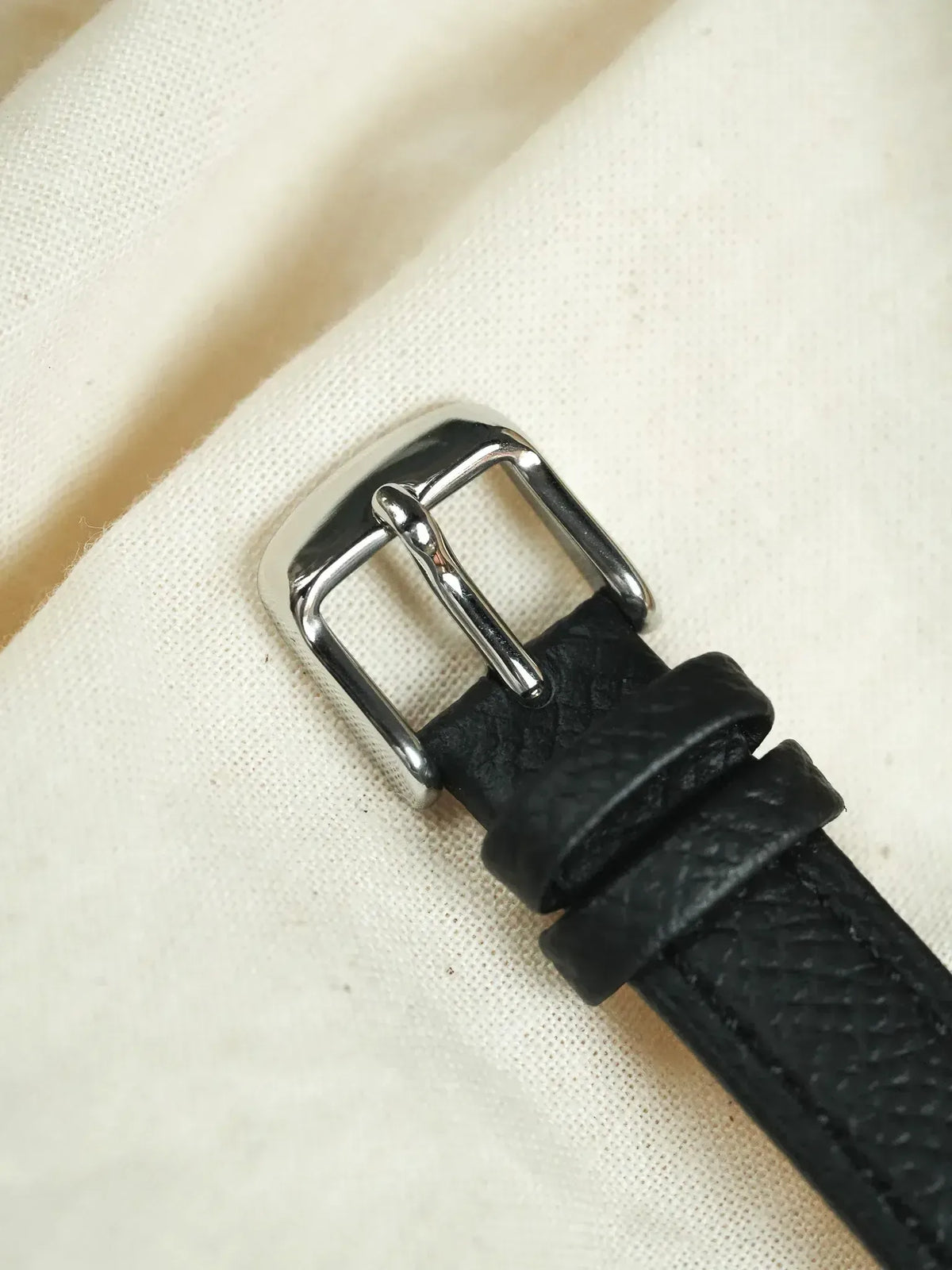 Black textured leather watch strap with polished silver buckle on cream fabric