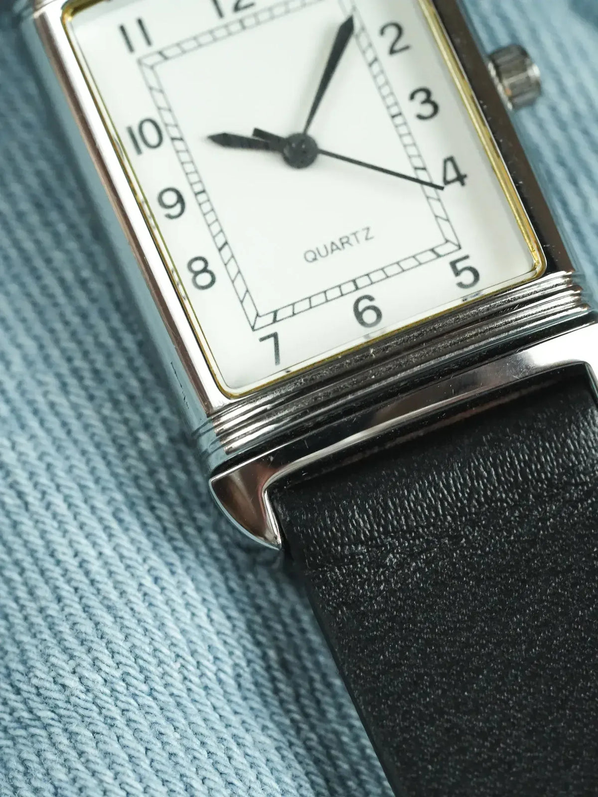 Vintage quartz watch with rectangular silver case, white dial, black leather strap on blue fabric
