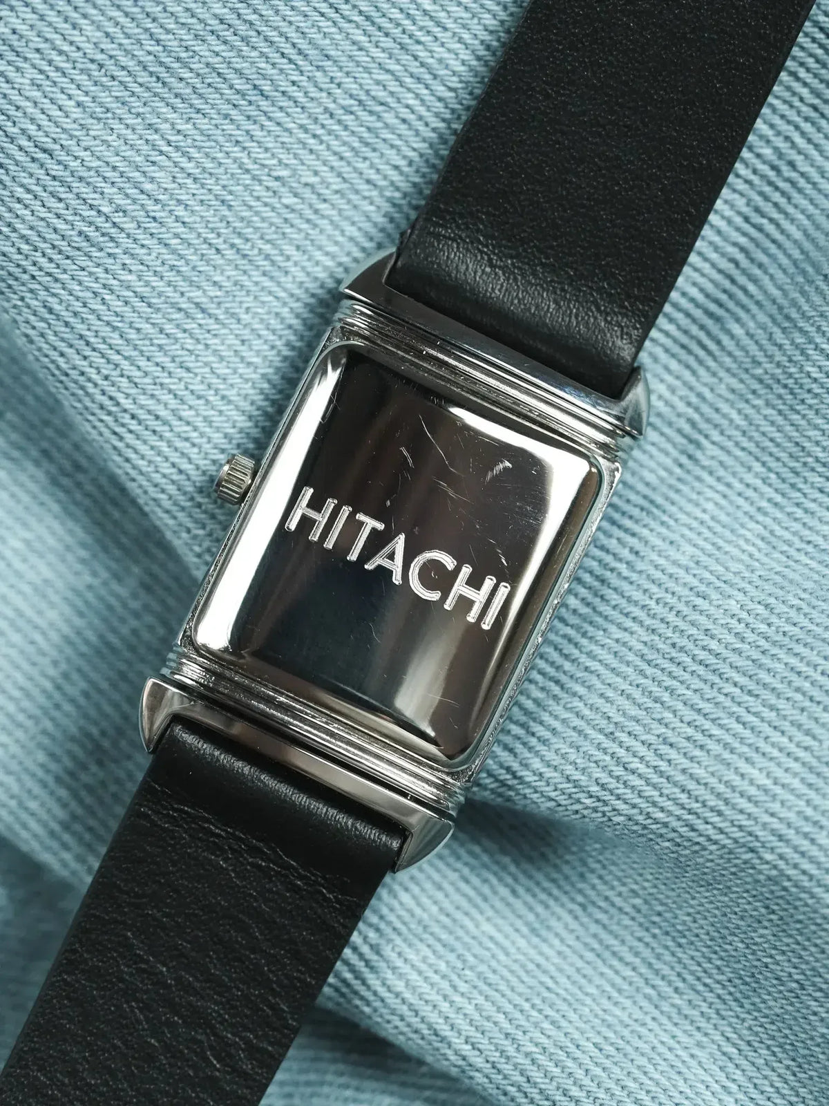 Hitachi vintage watch with rectangular silver case and black leather strap on blue fabric