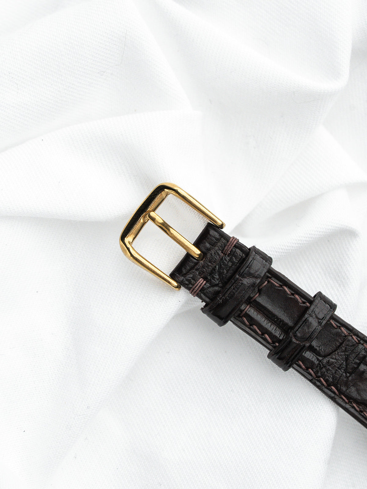 Boucle de montre dorée et bracelet en cuir brun façon alligator sur fond blanc