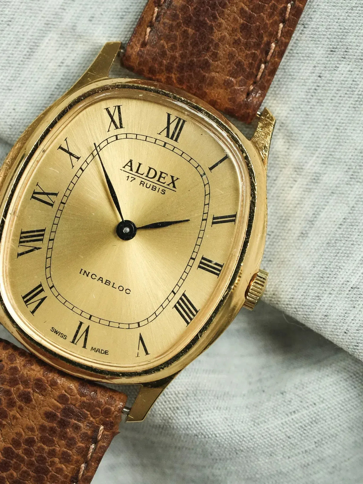 Aldex vintage watch with gold case, Roman numerals, brown leather strap on fabric