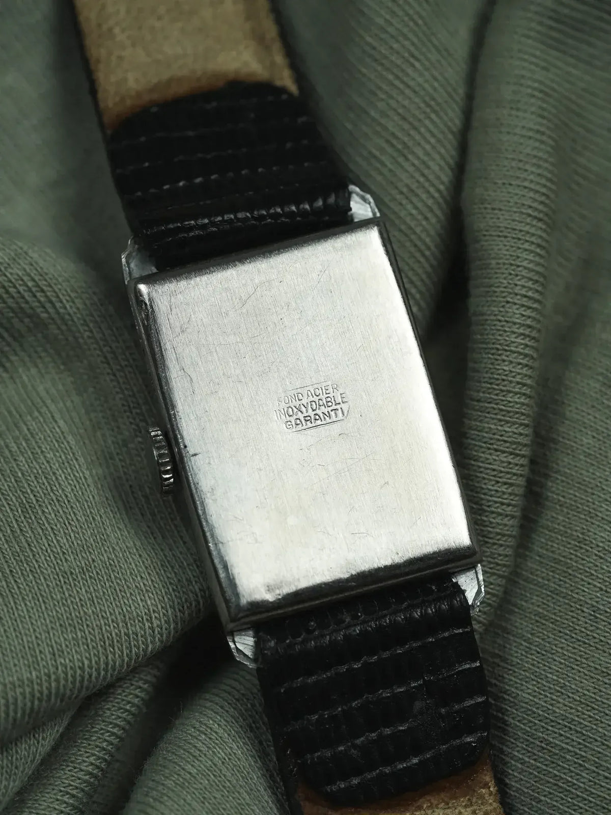 Vintage rectangular watch case back with stainless steel inscription, black leather strap on green fabric