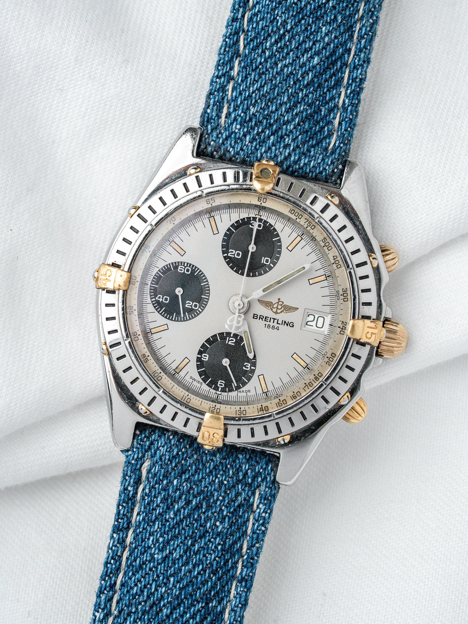 Breitling chronograph watch with silver dial, blue denim strap, gold accents on white background