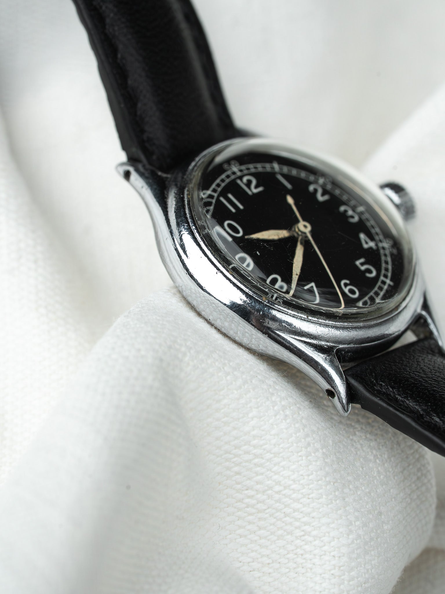 Vintage wristwatch with black dial, silver case, and black leather strap on white fabric