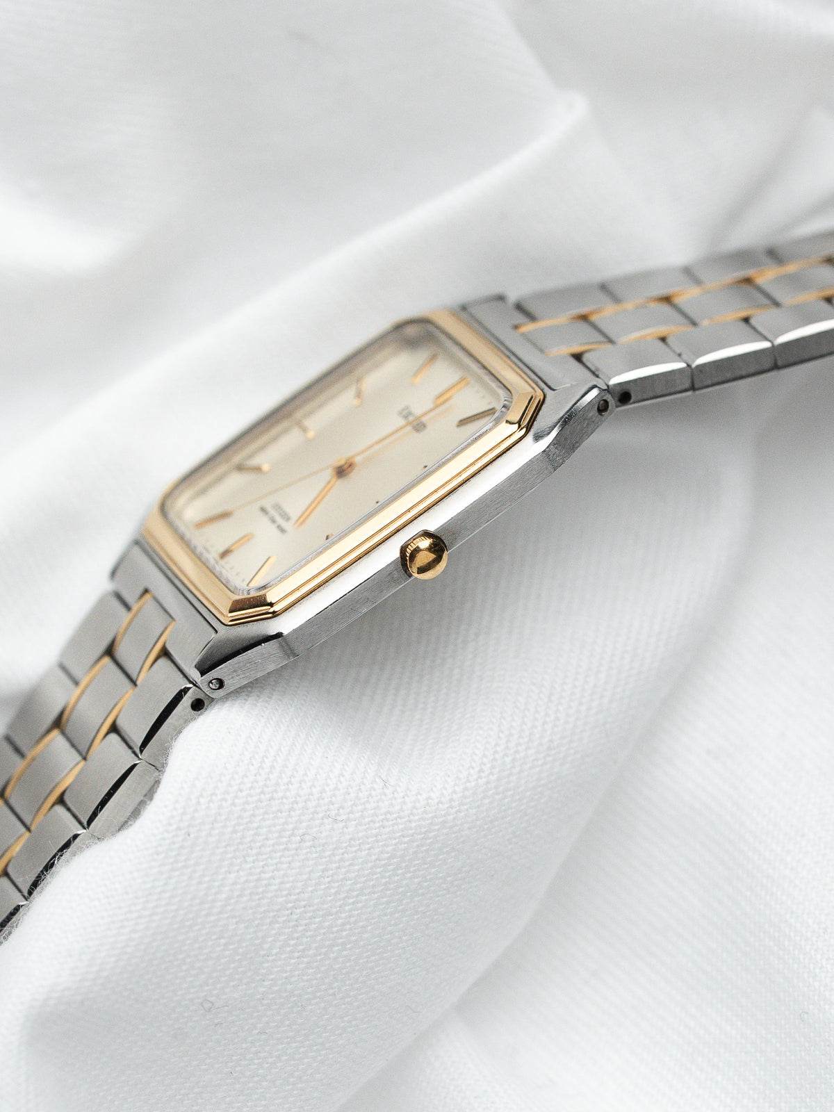 Montre vintage rectangulaire en acier et or avec cadran clair sur tissu blanc.