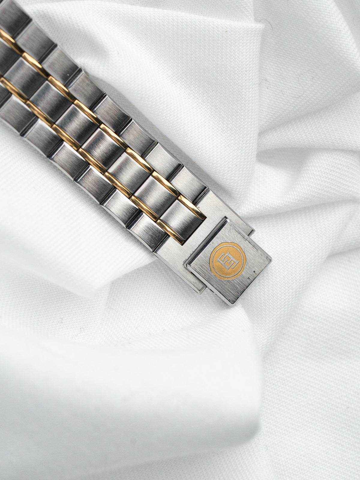 Détail d'un bracelet de montre en acier et or sur un tissu blanc.