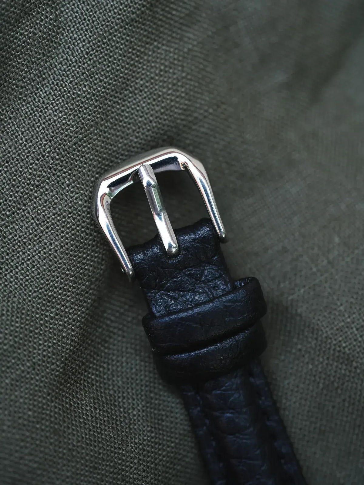 Black textured leather watch strap with polished steel buckle on fabric background