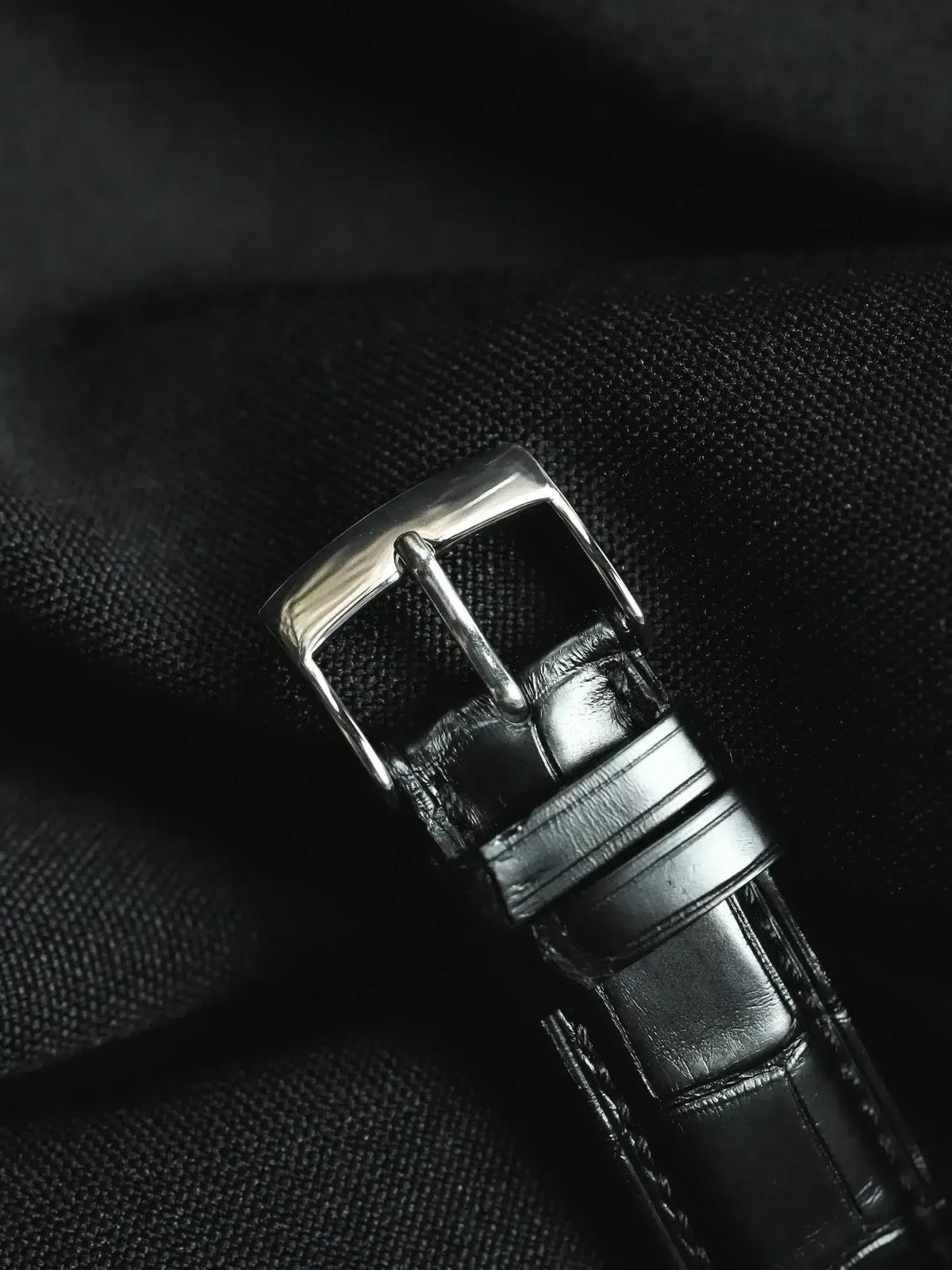 Black alligator leather watch strap with polished steel buckle on dark fabric
