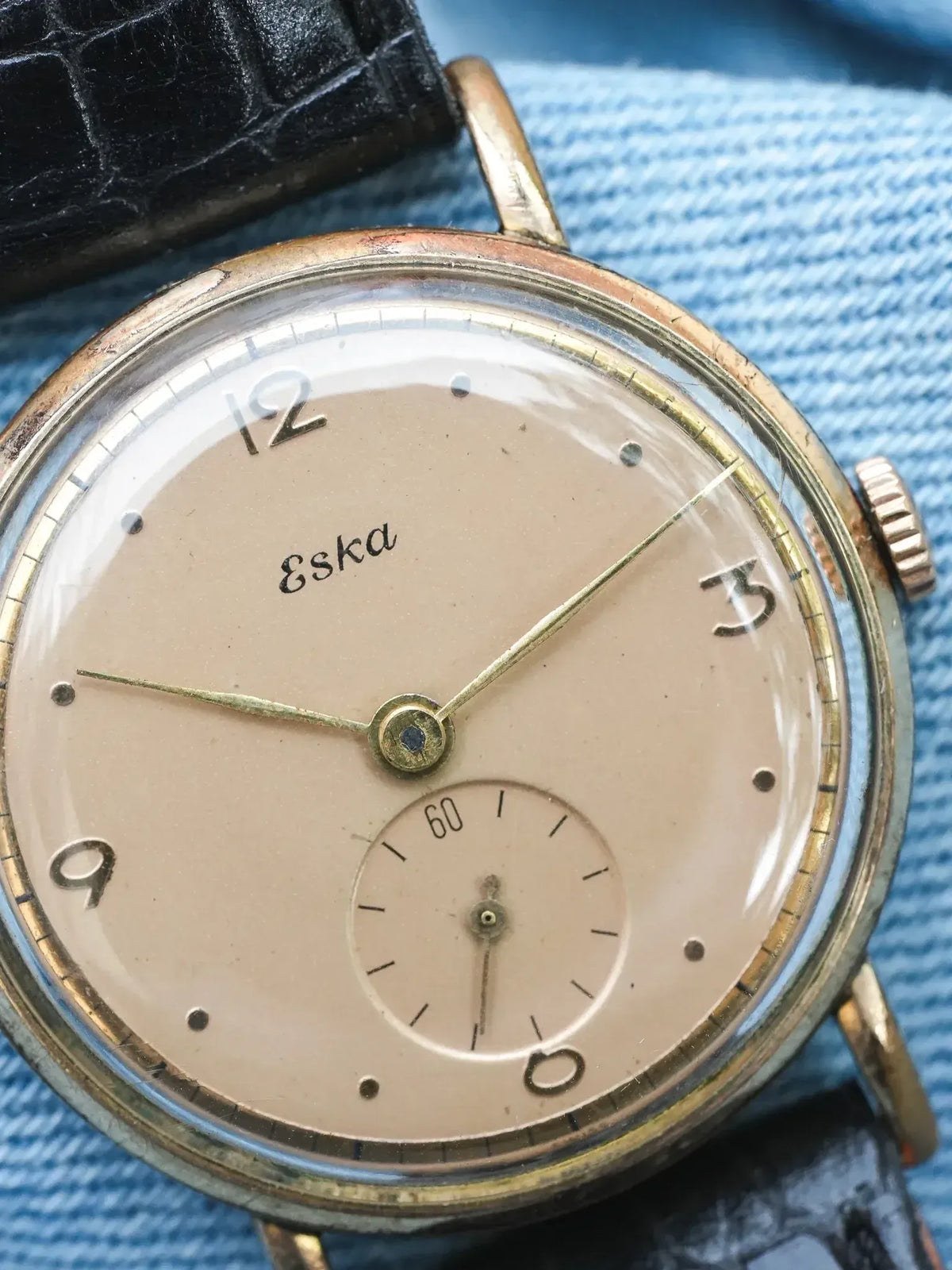 Vintage Eska wristwatch with cream dial, gold hands, black leather strap on denim