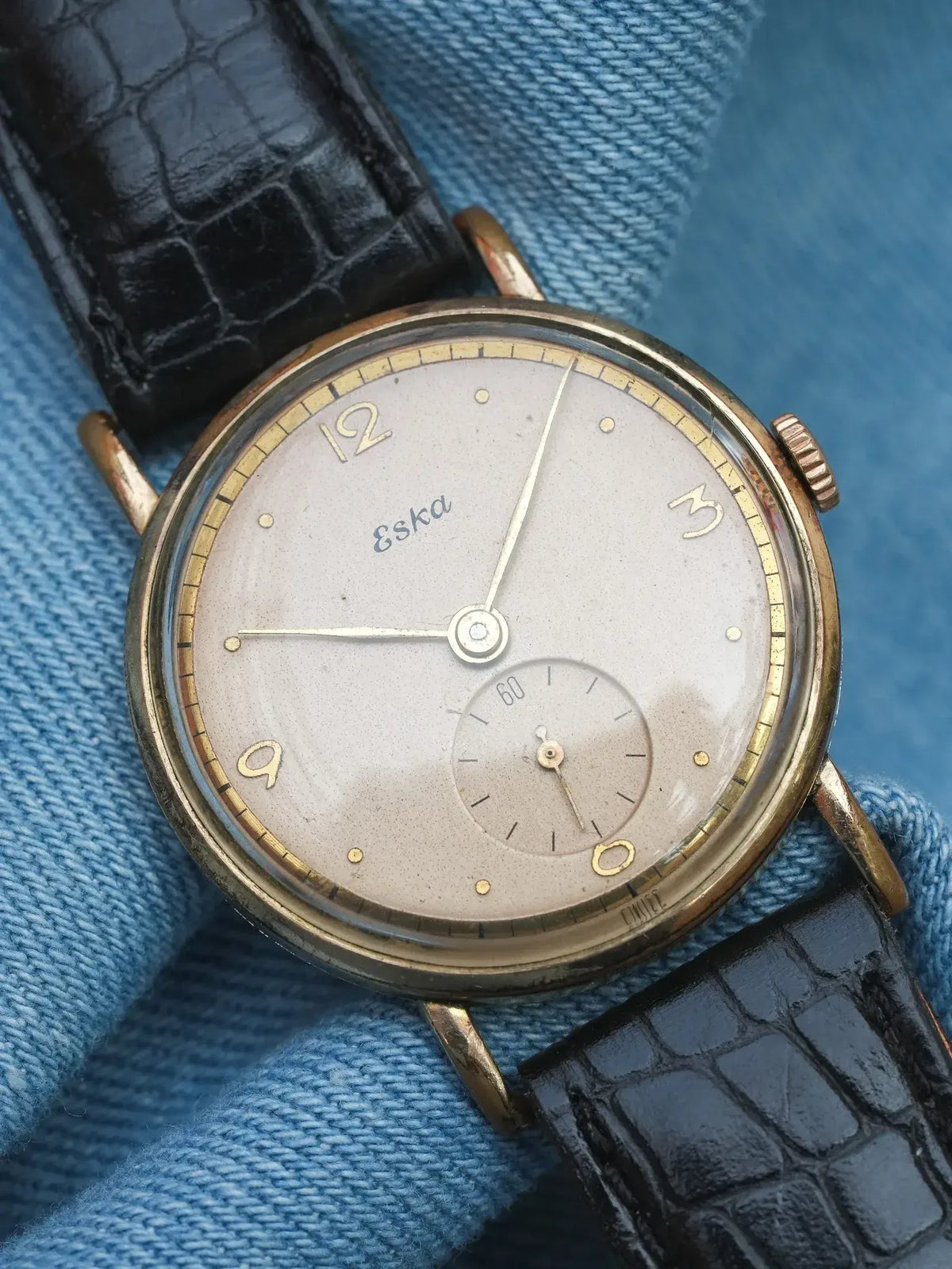 Vintage Eska wristwatch with gold-toned case, silvered dial, and black alligator strap on denim