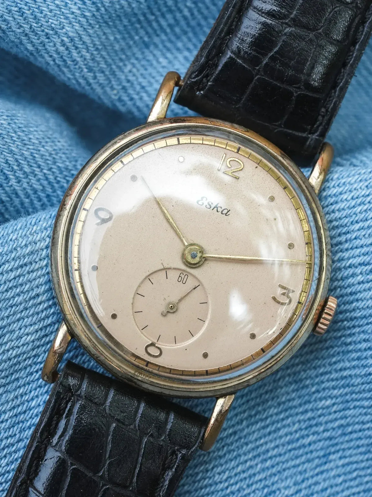 Vintage Eska wristwatch with cream dial, gold hands, and black crocodile strap on denim