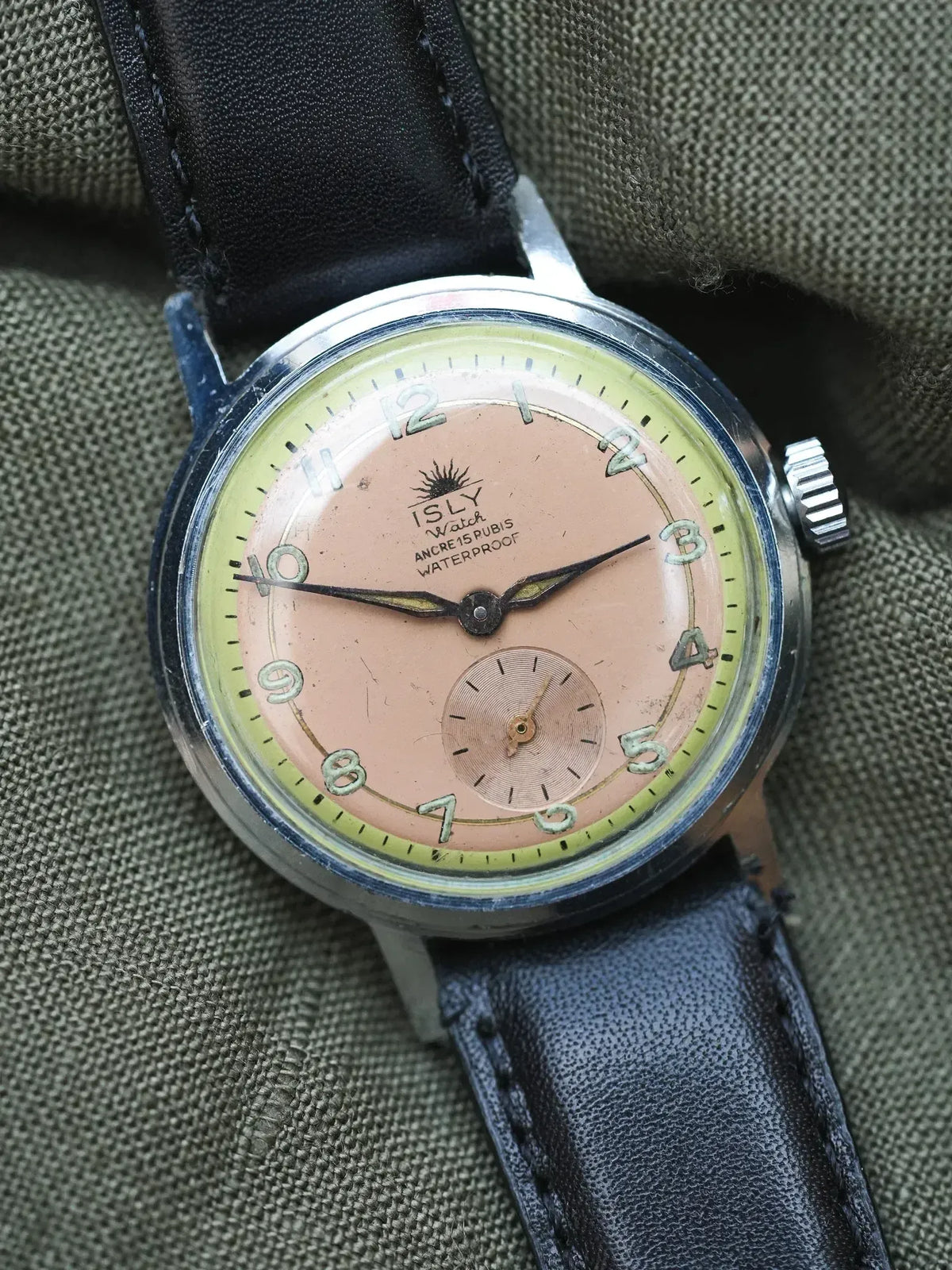 Vintage ISLY Watch with pink dial, black leather strap, resting on olive fabric