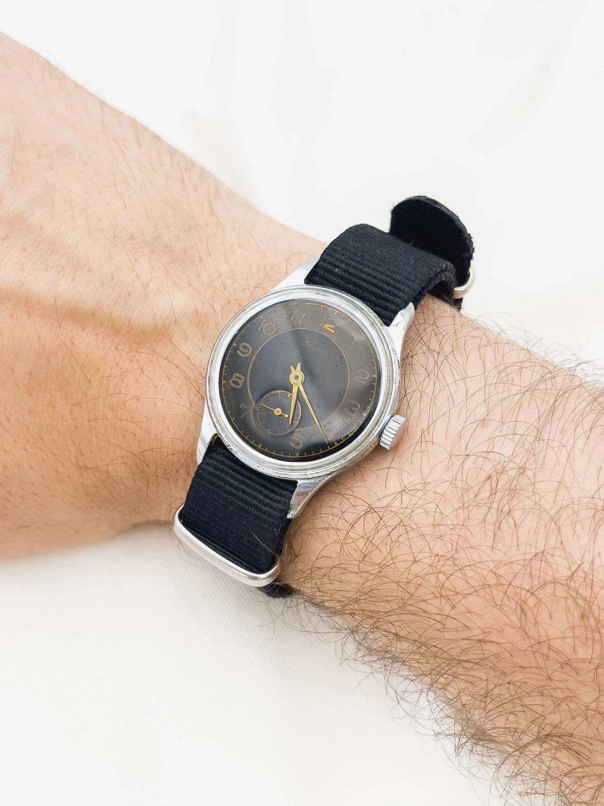 Vintage watch with black dial, gold hands, steel case, black fabric strap on wrist