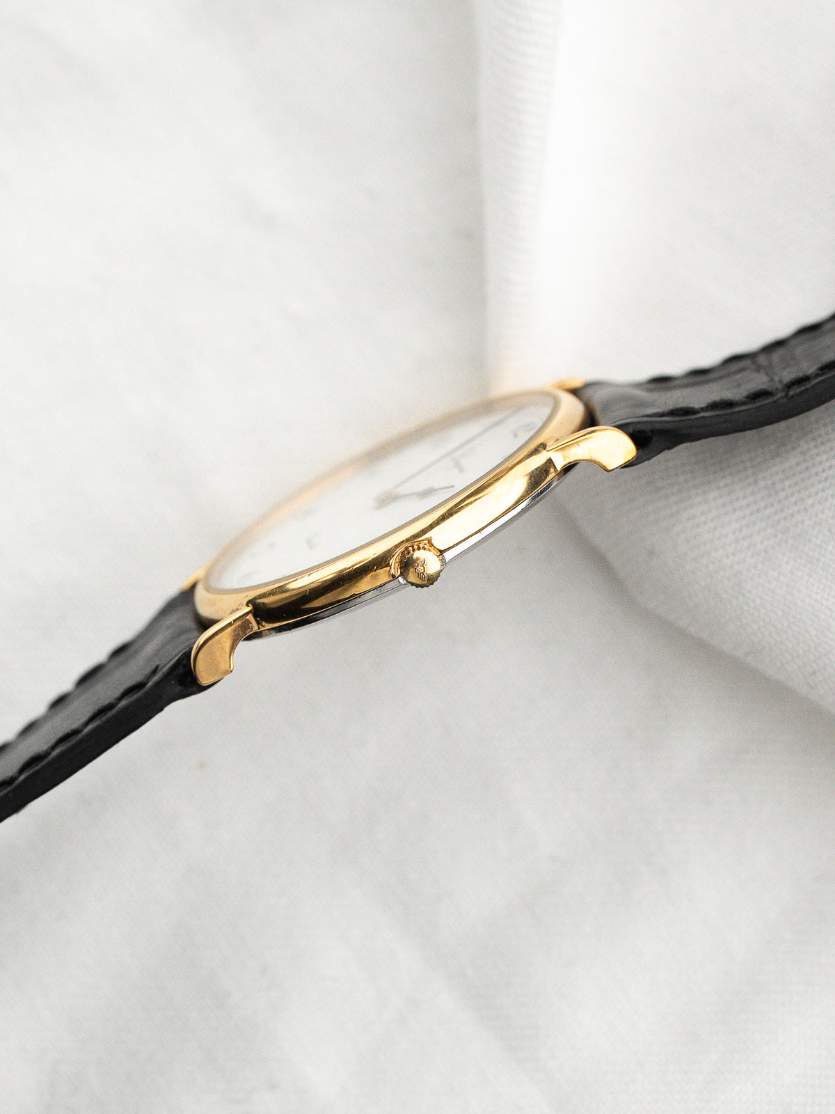 Montre vintage avec boîtier doré, cadran blanc et bracelet en cuir noir.