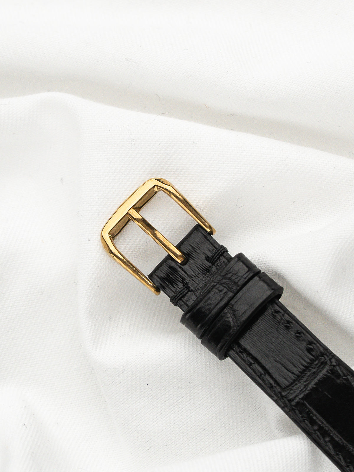 Boucle dorée et bracelet en cuir noir grainé sur un tissu blanc.