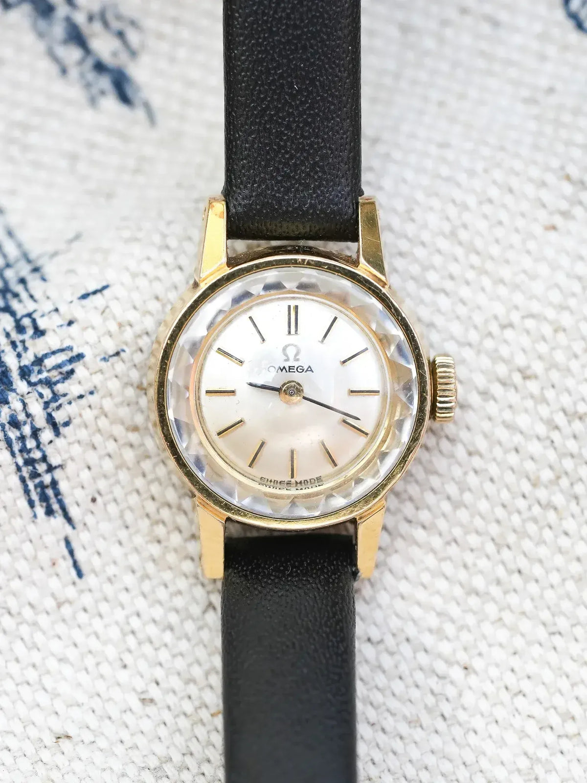 Vintage Omega watch with gold case, white dial, black leather strap on textured fabric