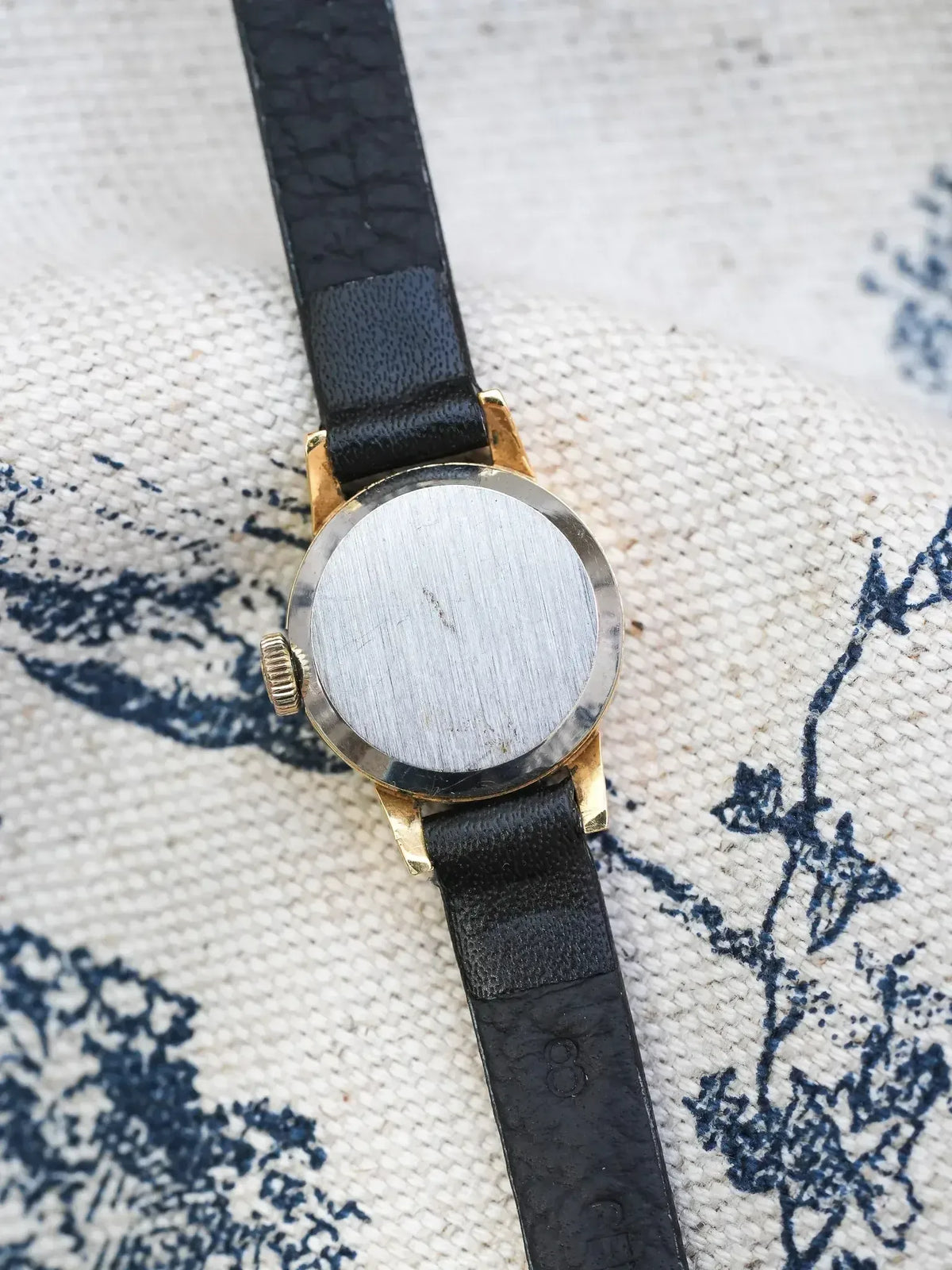Vintage watch with gold case, black leather strap, displayed on patterned fabric