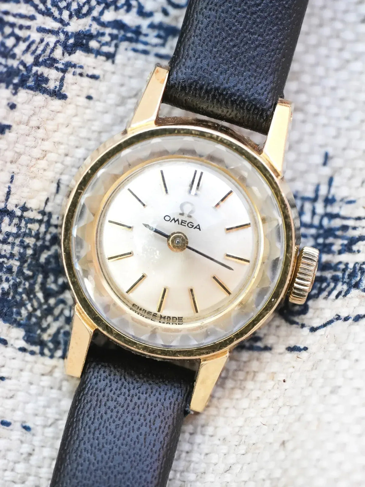 Vintage Omega watch with gold case, white dial, black leather strap on textured fabric