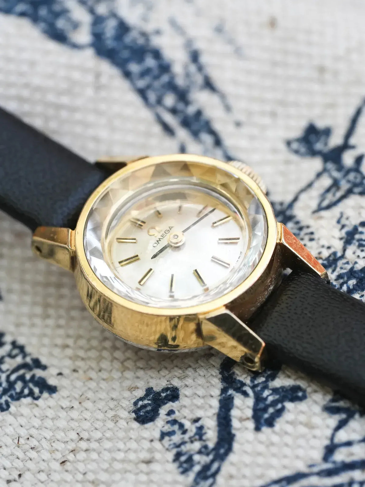 Vintage Omega watch with gold case, white dial, black leather strap on patterned fabric