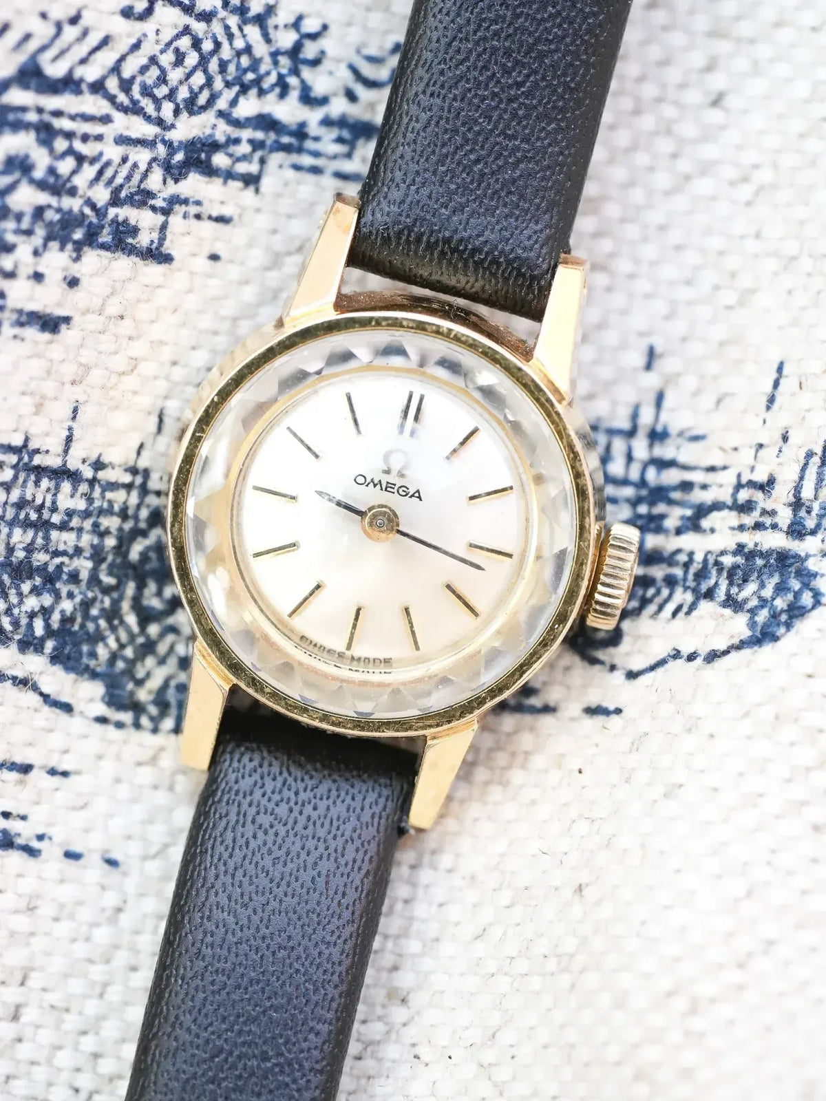 Vintage Omega watch with gold case, white dial, black leather strap on patterned fabric