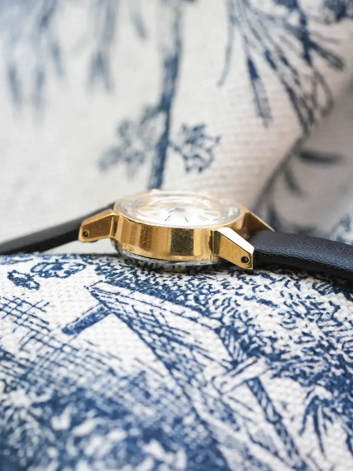 Vintage gold watch with black leather strap on blue white patterned fabric
