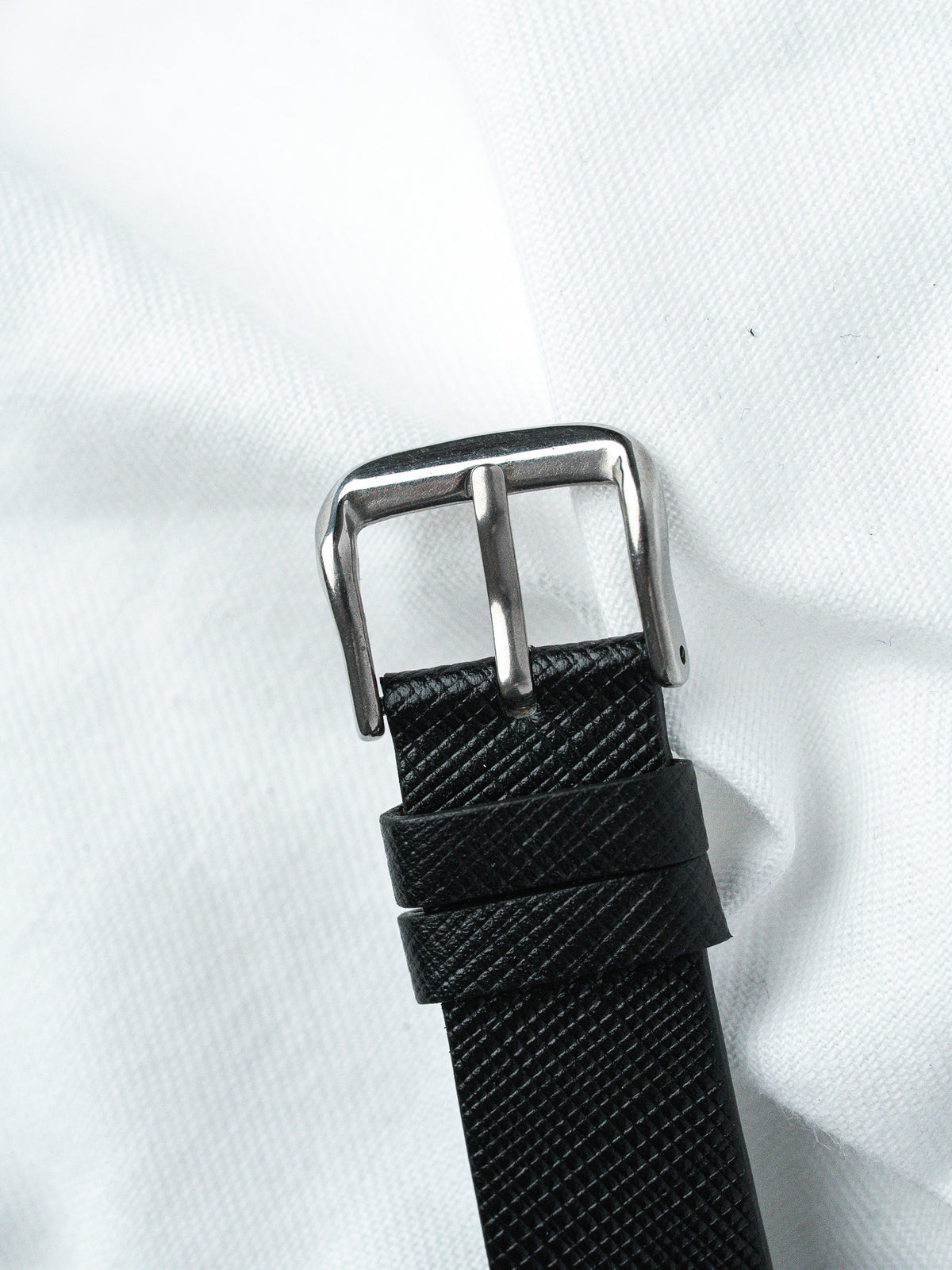 Black textured leather watch strap with polished steel buckle on white fabric