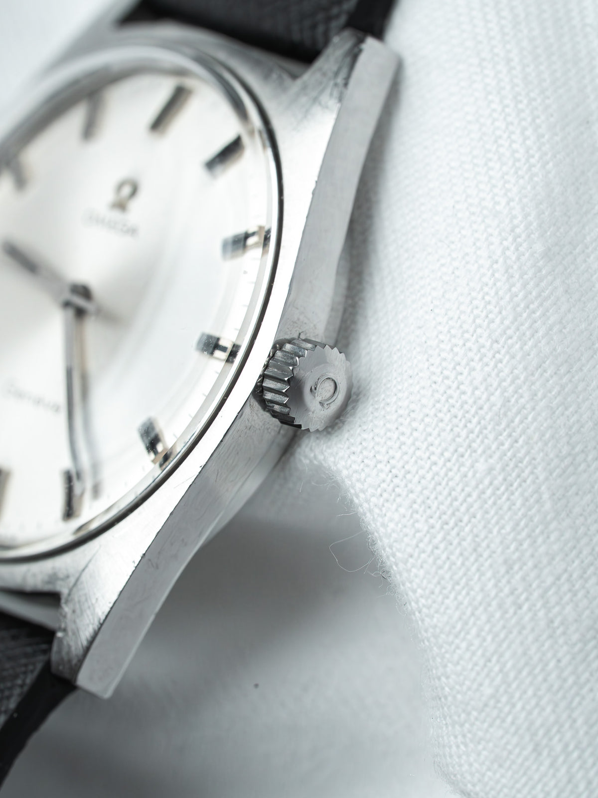 Vintage Omega watch with silver dial, black strap, and textured crown on white fabric
