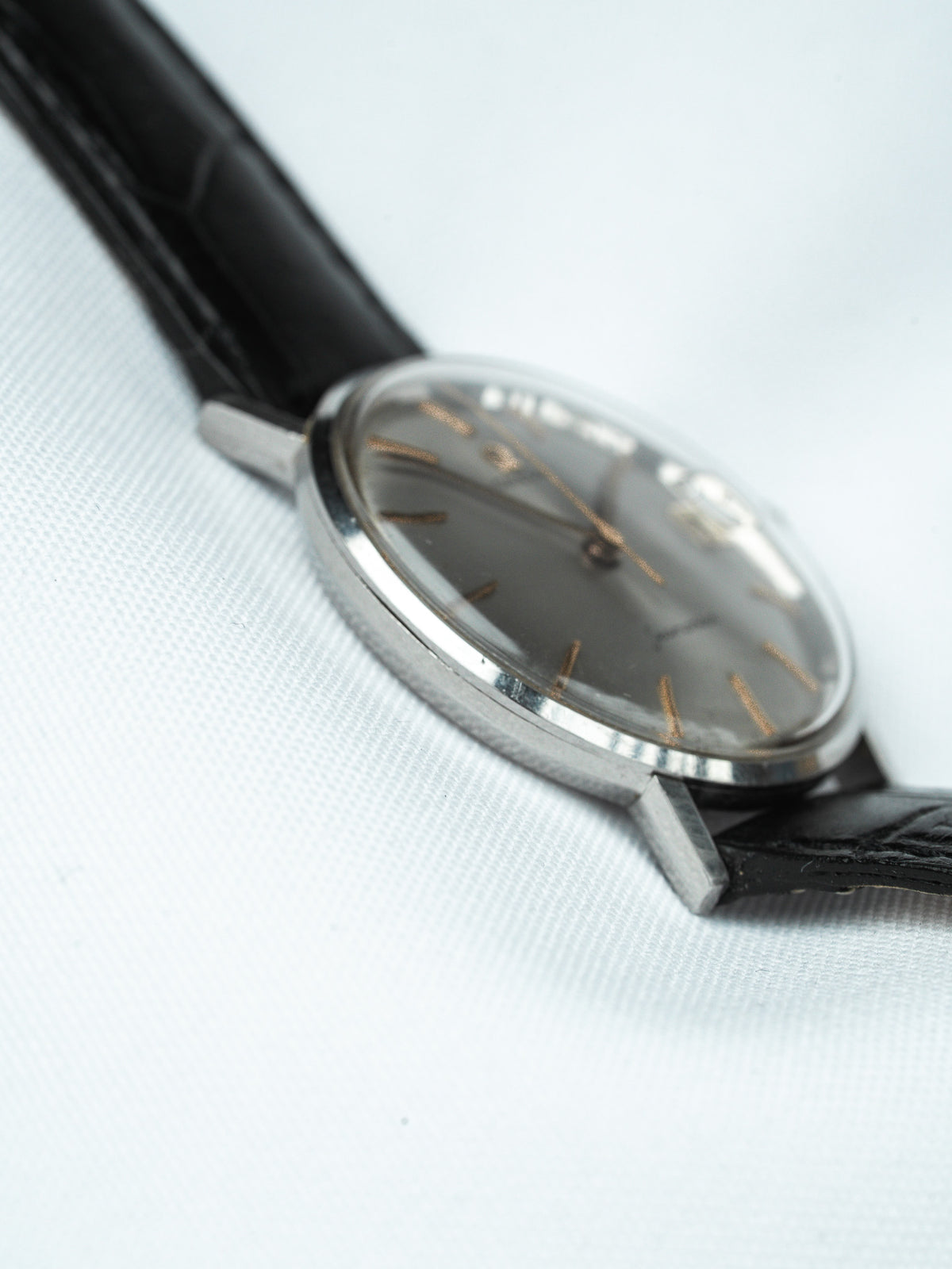 Vintage Universal Genève watch with grey dial, gold markers, steel case, black leather strap on white fabric