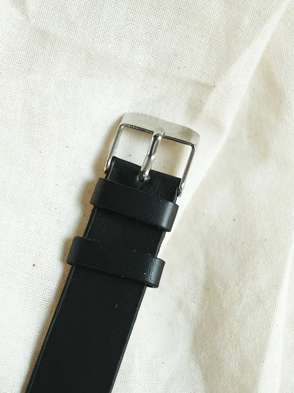 Black leather watch strap with silver buckle on white fabric background