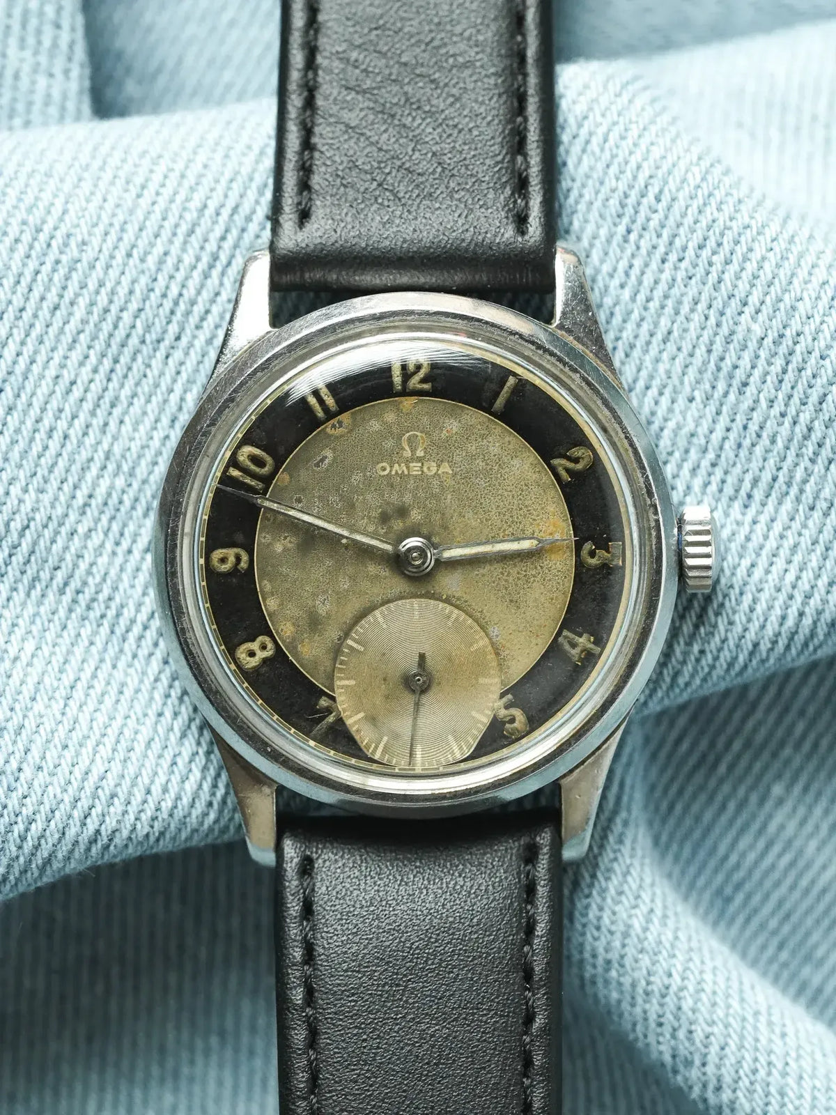 Vintage Omega wristwatch with aged dial, black leather strap, on denim background