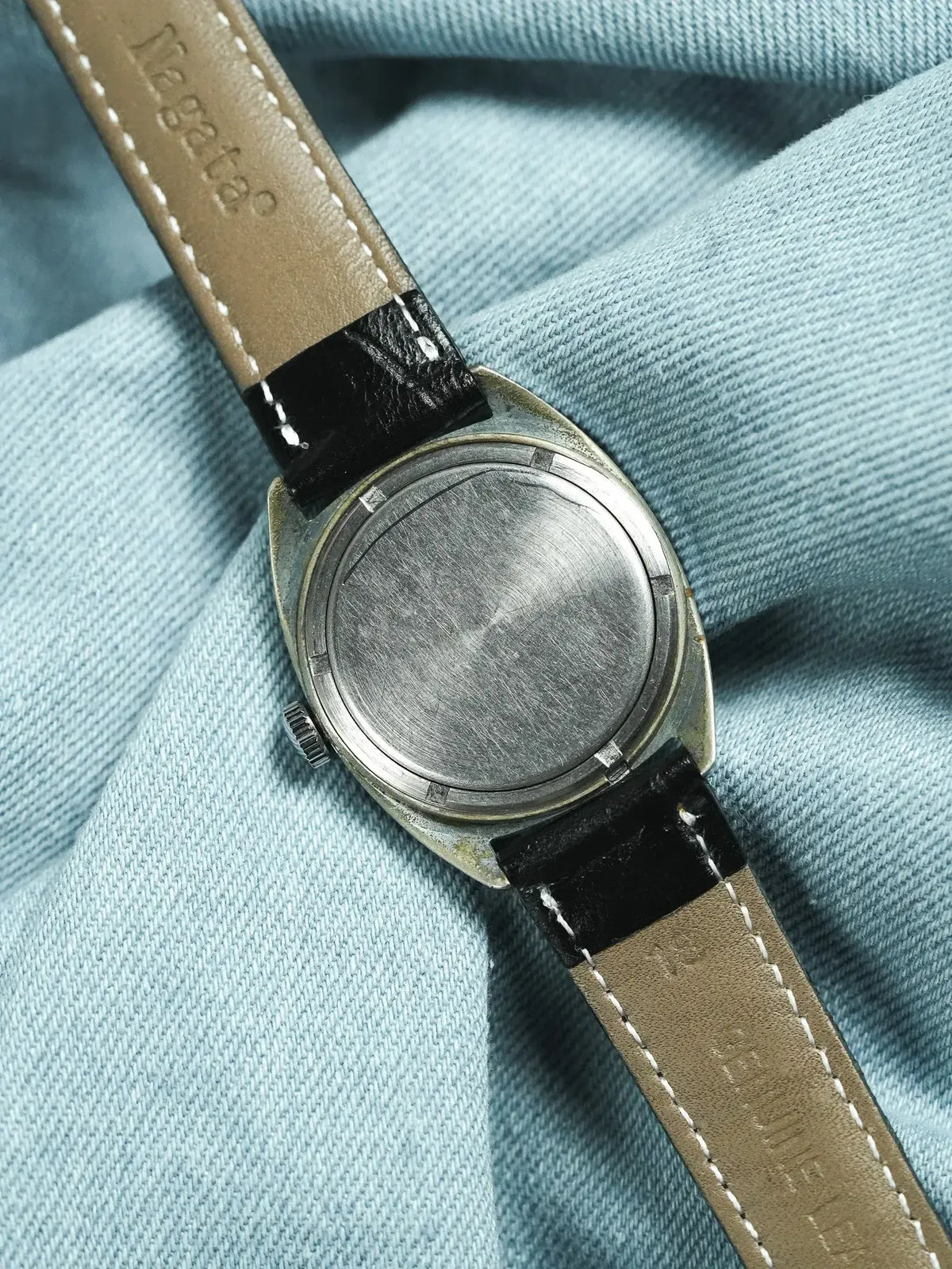 Vintage watch with brushed steel case back, tan leather strap, resting on blue fabric