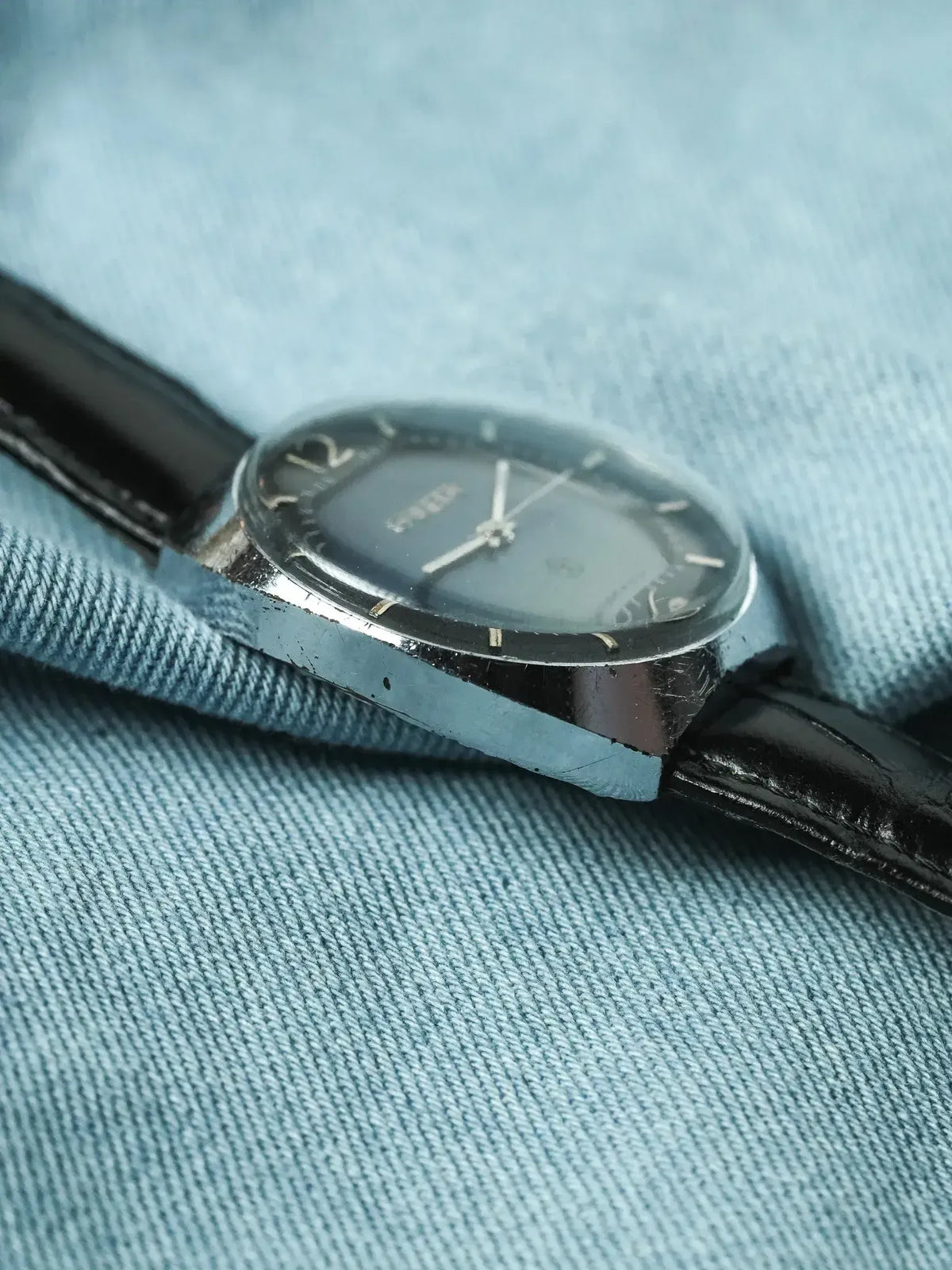 Vintage Omega watch with black dial, steel case, and black leather strap on blue fabric