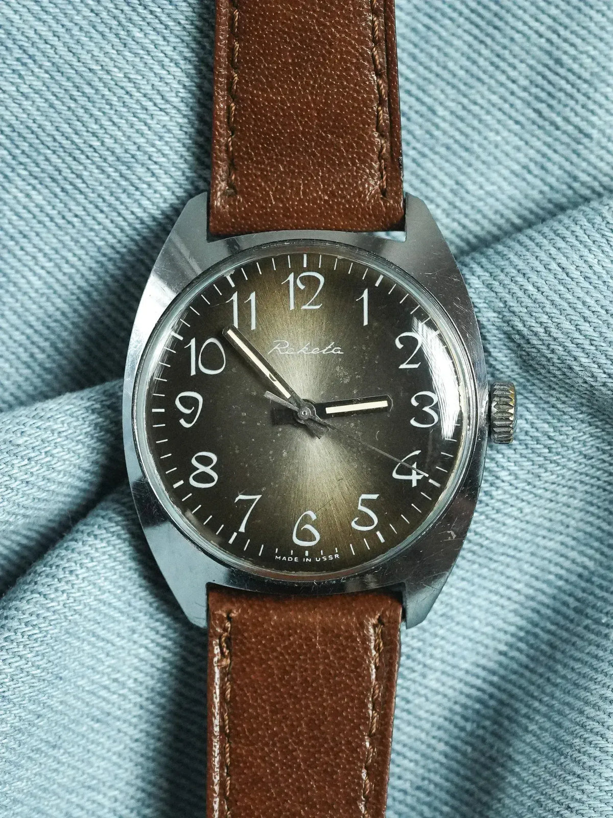 Raketa vintage watch with black dial, white numerals, brown leather strap on denim