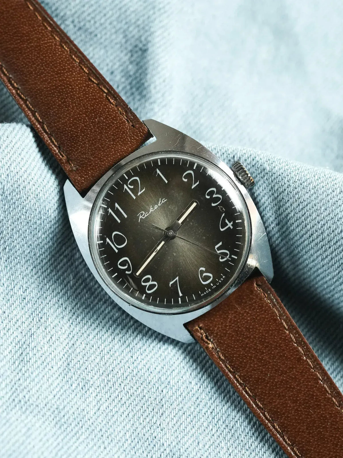 Raketa vintage watch with black dial, white numerals, brown leather strap on denim