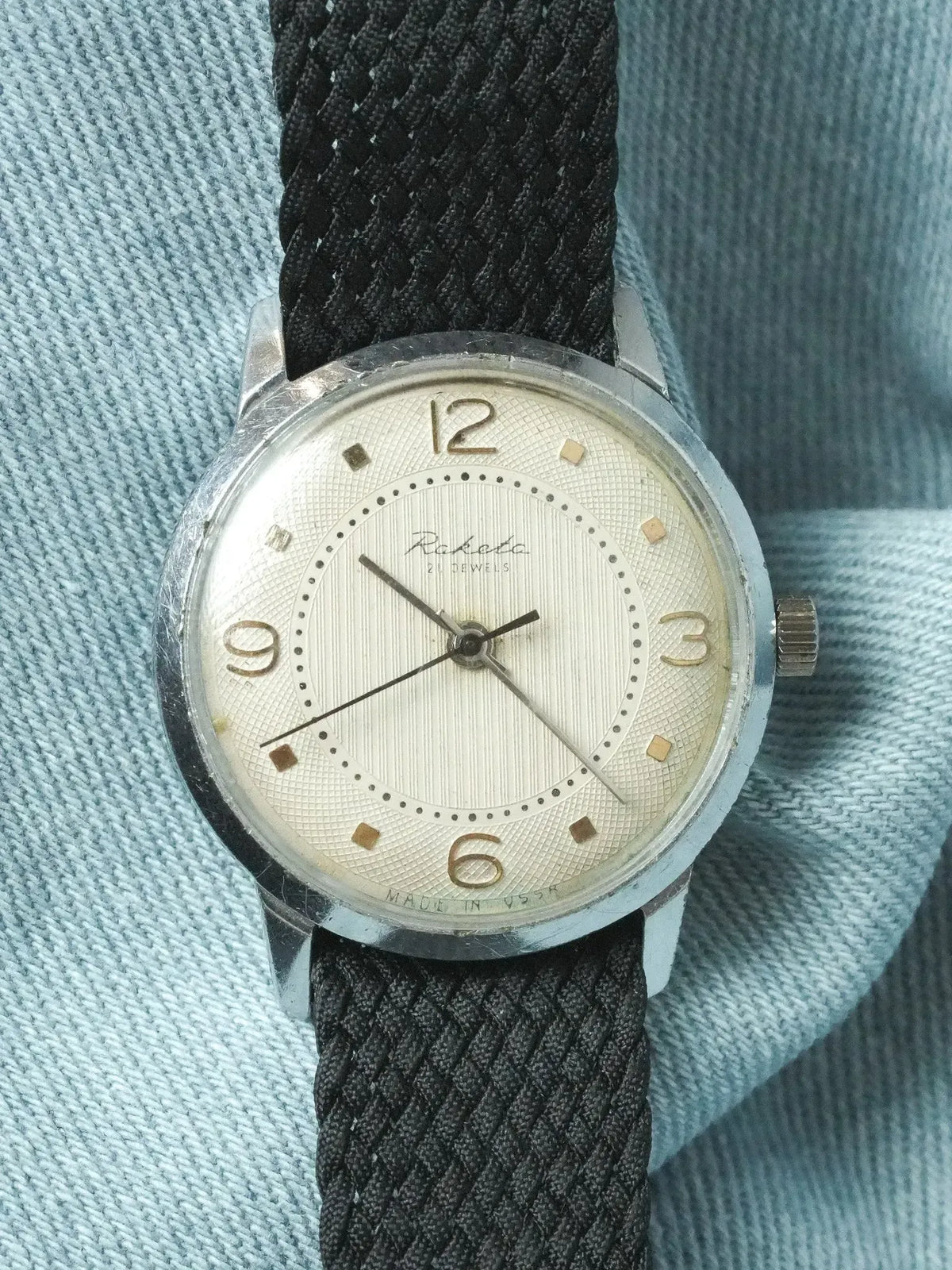 Vintage Raketa watch with silver dial, black fabric strap, worn on denim